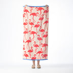 Pink beach towel featuring a repeating coral flamingo design with contrasting blue borders, held up against a white background.