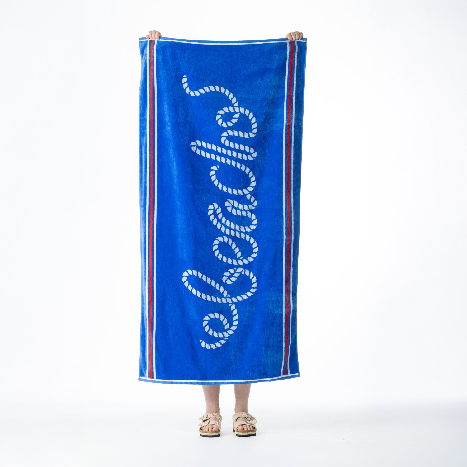 Blue beach towel featuring a white rope-style "Beach" design with contrasting red and white striped borders, held up against a white background.