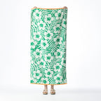Green tropical beach towel featuring a repeating hibiscus flower and palm leaf design with contrasting orange borders, held up against a white background.