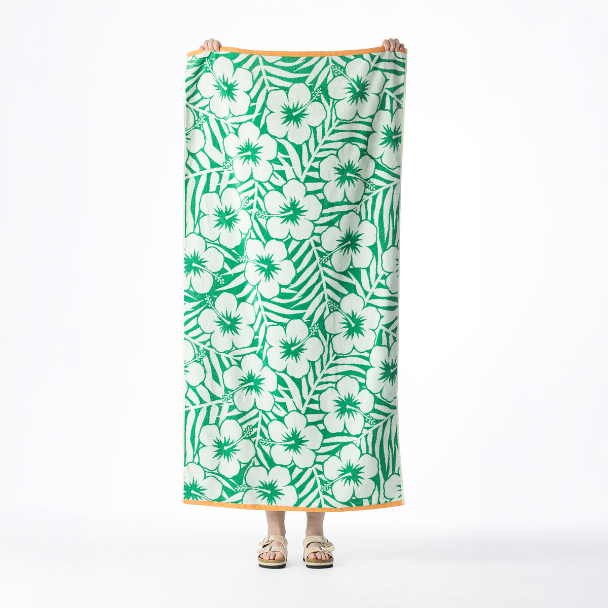 Green tropical beach towel featuring a repeating hibiscus flower and palm leaf design with contrasting orange borders, held up against a white background.