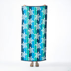 Multicoloured beach towel featuring a repeating white turtle design over vertical blue, aqua and teal stripes, held up against a white background.