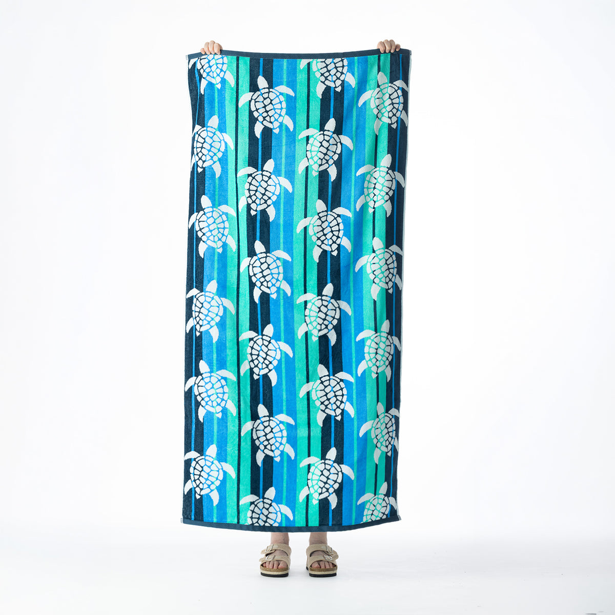 Multicoloured beach towel featuring a repeating white turtle design over vertical blue, aqua and teal stripes, held up against a white background.
