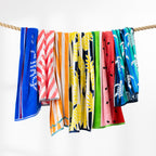 Colourful beach towels featuring assorted tropical and summer designs draped over a rope against a white background.