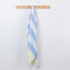 A striped beach towel with light blue, white, and pale yellow accents hangs from a light wood peg rack against a plain white wall.