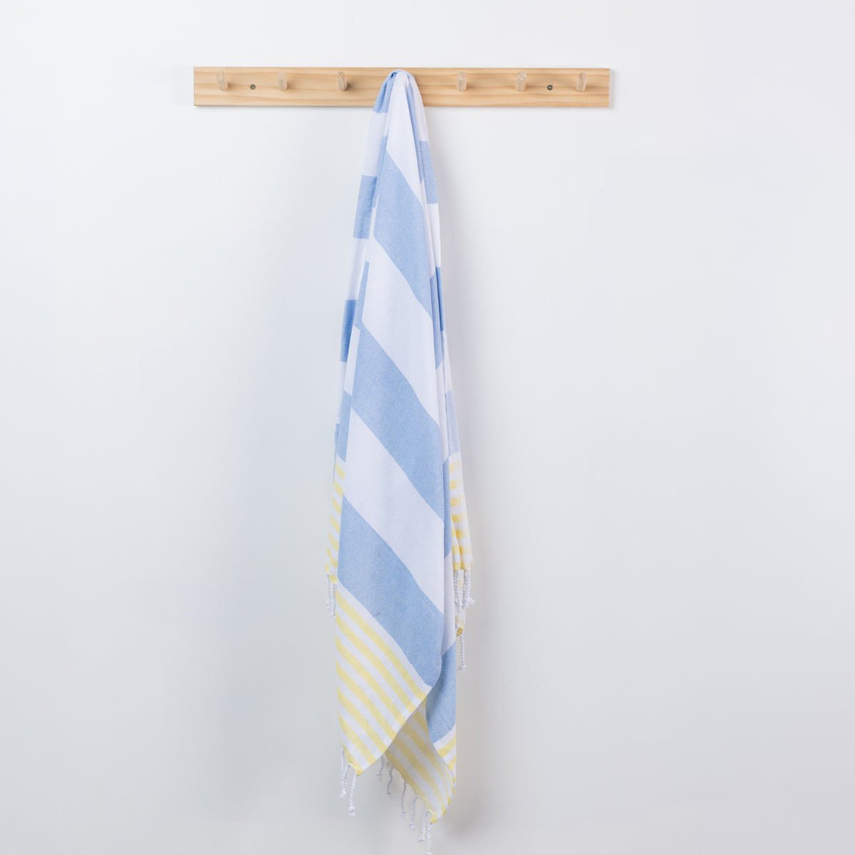 A striped beach towel with light blue, white, and pale yellow accents hangs from a light wood peg rack against a plain white wall.