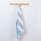 A striped beach towel with alternating light gray and white stripes, and teal stripes along one edge, hanging from a light wood peg on a white wall.