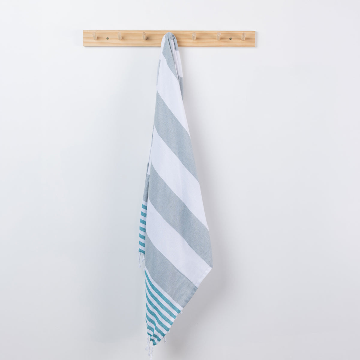 A striped beach towel with alternating light gray and white stripes, and teal stripes along one edge, hanging from a light wood peg on a white wall.