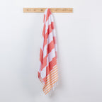 A striped beach towel with white and coral-red diagonal stripes and orange-and-white striped fringe, hanging from a light wood peg rail against a plain white wall.