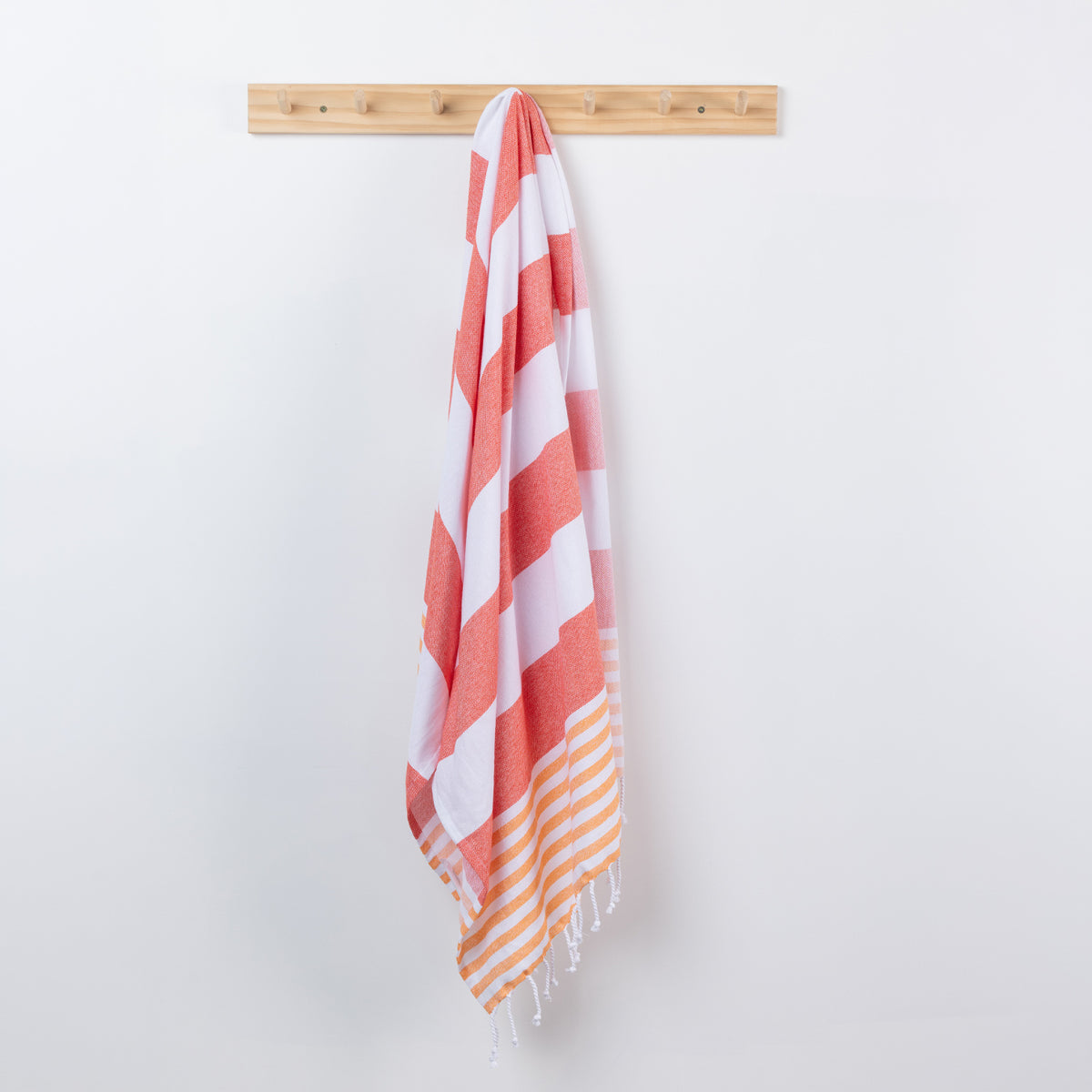 A striped beach towel with white and coral-red diagonal stripes and orange-and-white striped fringe, hanging from a light wood peg rail against a plain white wall.