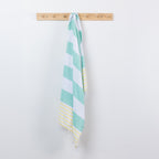 A seafoam green, white, and yellow striped cotton towel with fringed edges hangs from a light wood peg on a plain white wall.