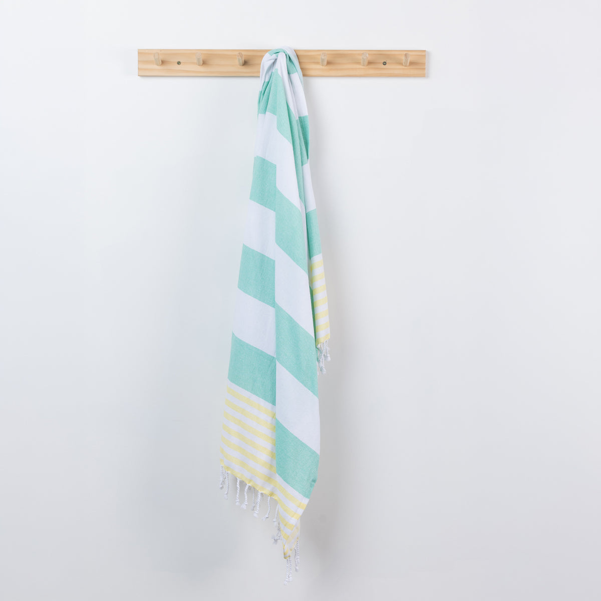 A seafoam green, white, and yellow striped cotton towel with fringed edges hangs from a light wood peg on a plain white wall.