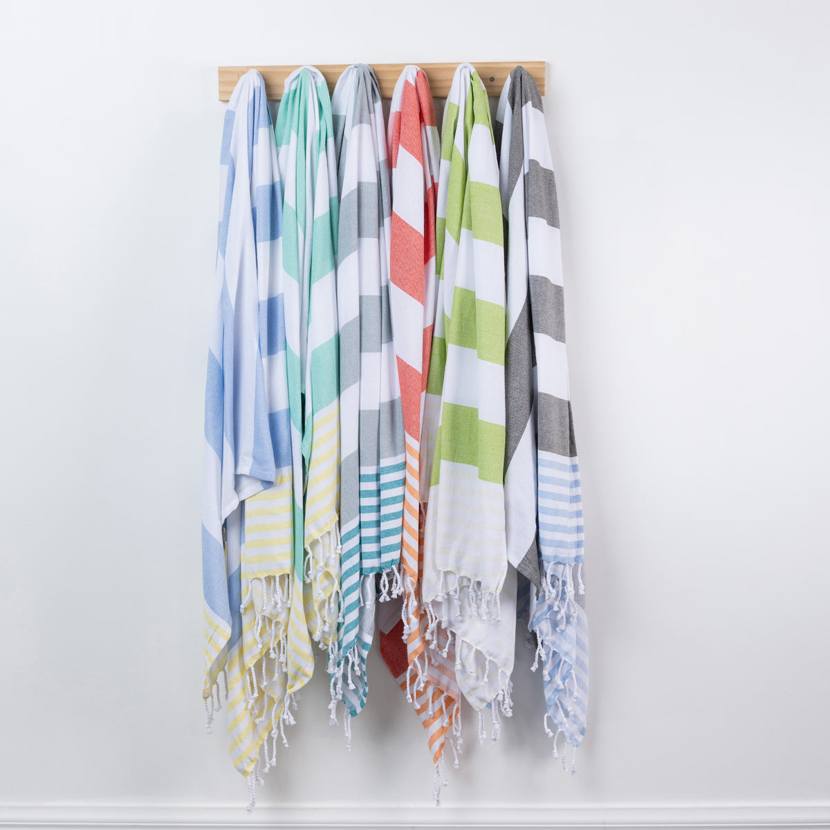 Six striped beach towels in assorted colors hang from a wooden rack against a plain white wall.