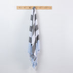 A striped beach towel with fringed edges hangs from a light wood peg rack against a plain white wall. The towel features alternating blocks of white, dark gray, and light blue, with a section of light blue and white stripes near the bottom.