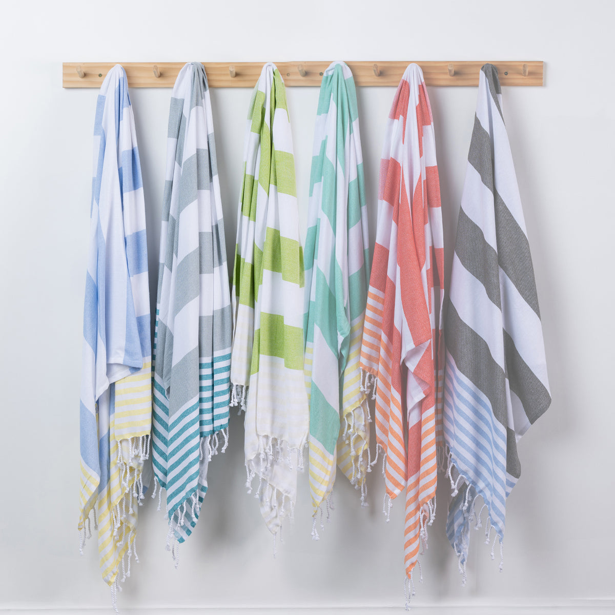 Six striped cotton beach towels in assorted colors hang from a wooden rack against a plain white wall.