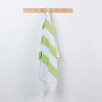 A white cotton beach towel with lime green diagonal stripes hangs from a light wood peg on a plain white wall.