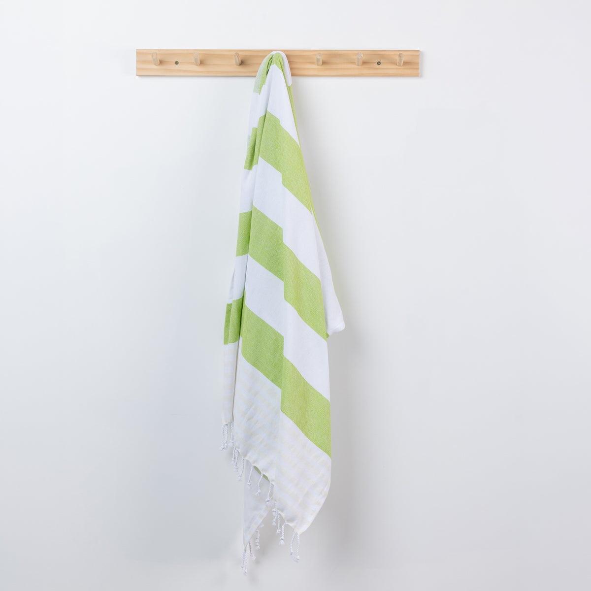 A white cotton beach towel with lime green diagonal stripes hangs from a light wood peg on a plain white wall.