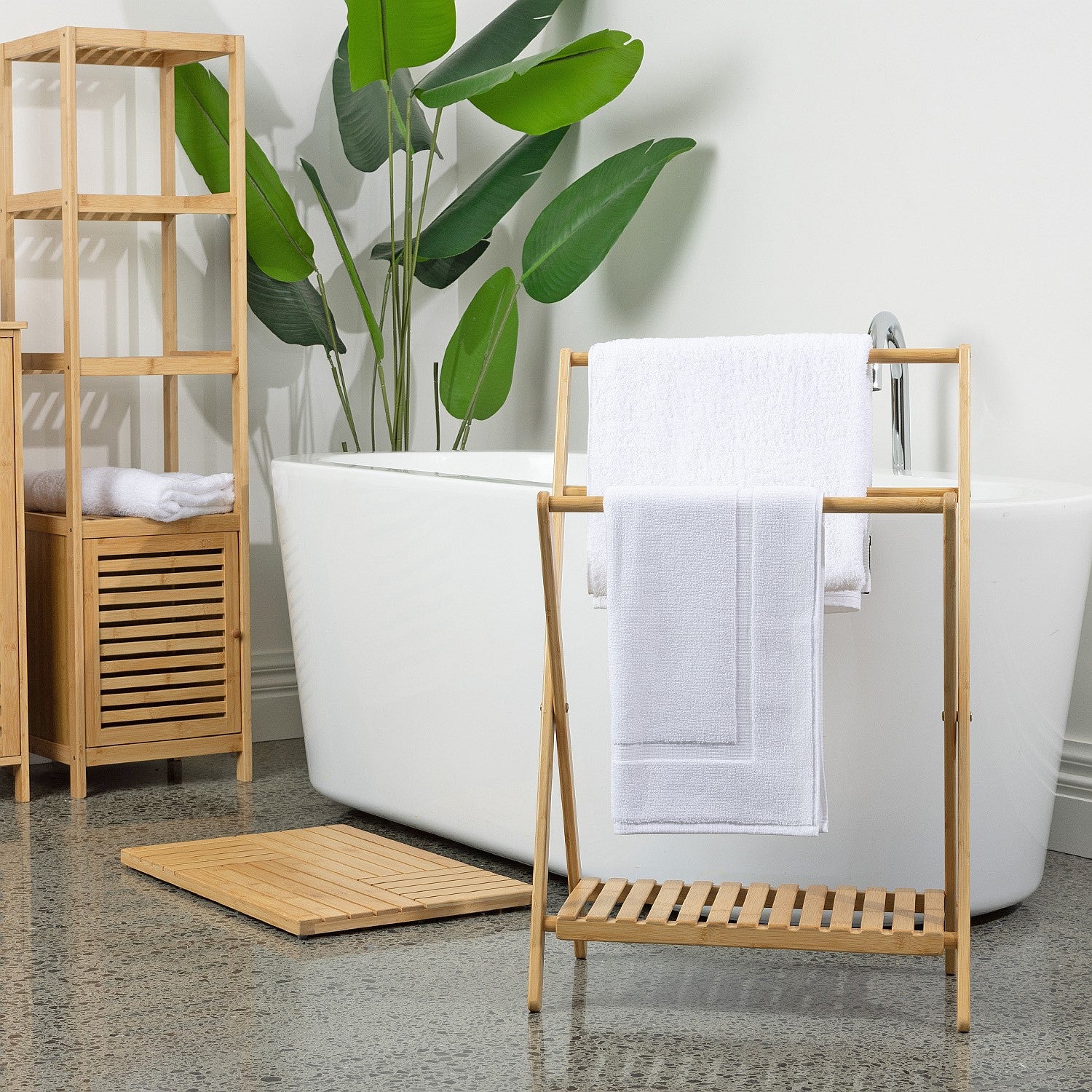 Bamboo bathroom accessories styled in a modern bathroom setting featuring a towel rack, shelving unit, bamboo bath mat, and freestanding bathtub with tropical greenery.
