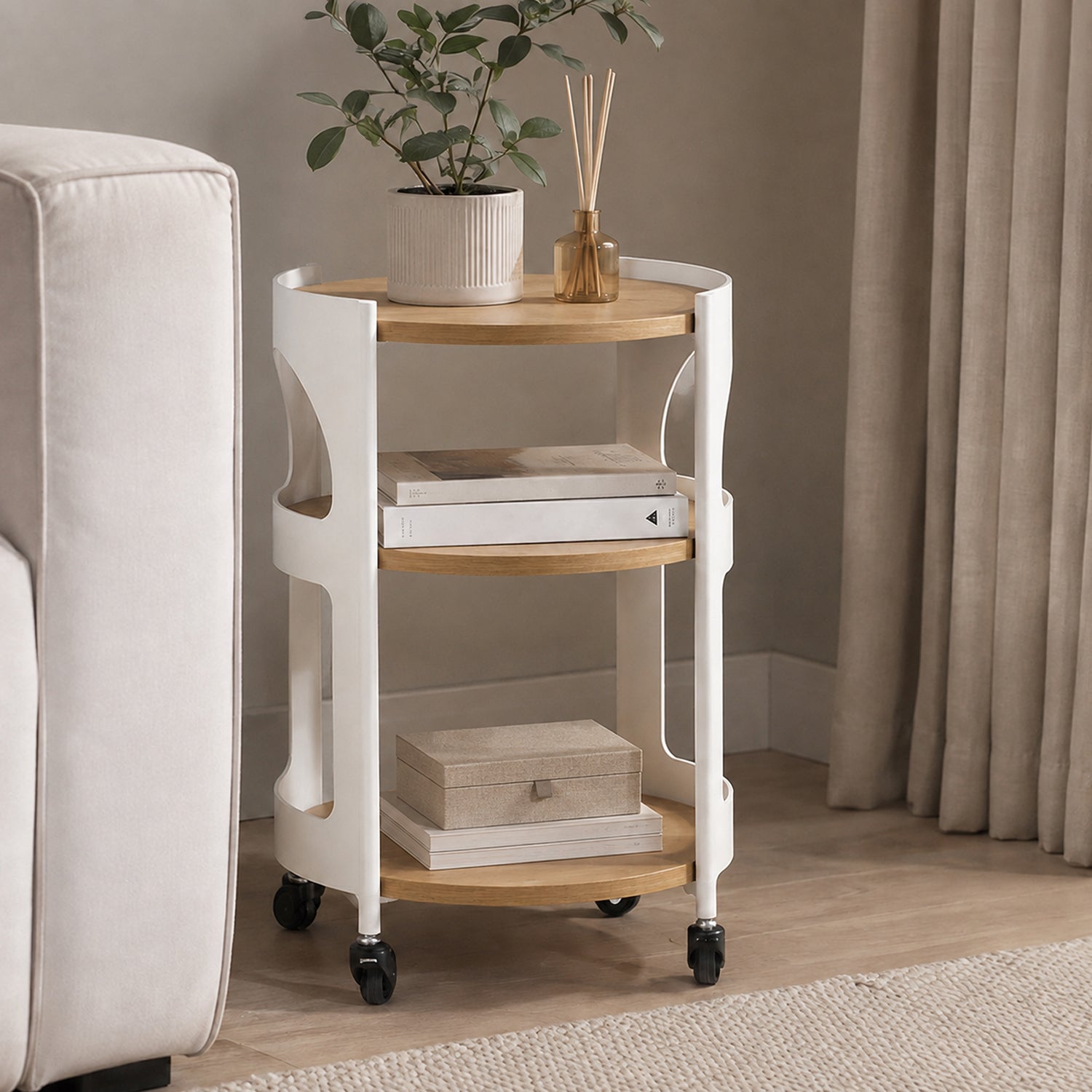 Solace Parker 3 Tier Trolley in white and natural styled beside a sofa, with a plant and diffuser on the top shelf, books on the middle shelf and a decorative box below. 