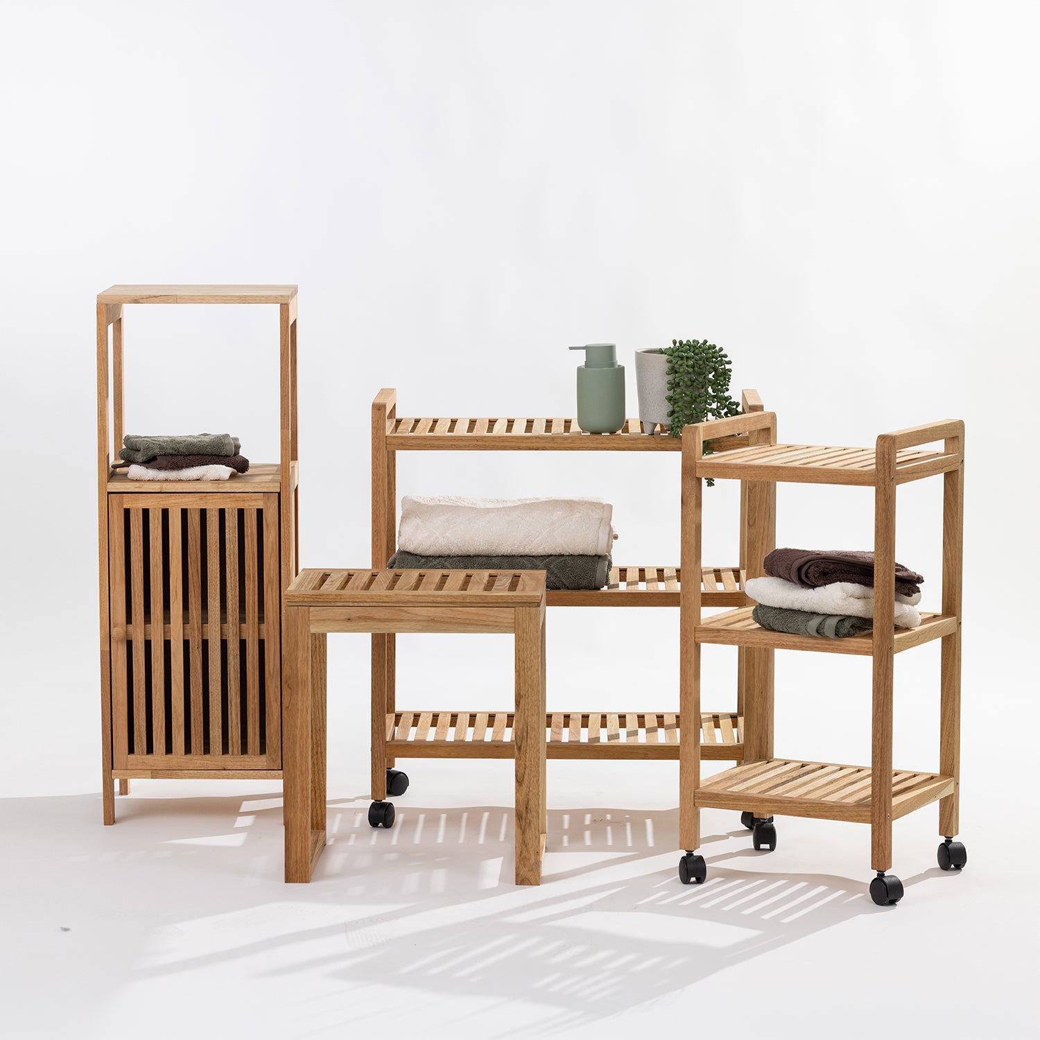 Three light natural wood bathroom storage organizers with slatted shelves and a slatted front cabinet door, shown on a clean white studio background. The set includes a tall narrow organizer, a medium shelving unit with folded towels and small decor items, and a rolling three-tier cart on black caster wheels.