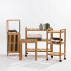 Three light natural wood bathroom storage organizers with slatted shelves and a slatted front cabinet door, shown on a clean white studio background. The set includes a tall narrow organizer, a medium shelving unit with folded towels and small decor items, and a rolling three-tier cart on black caster wheels.