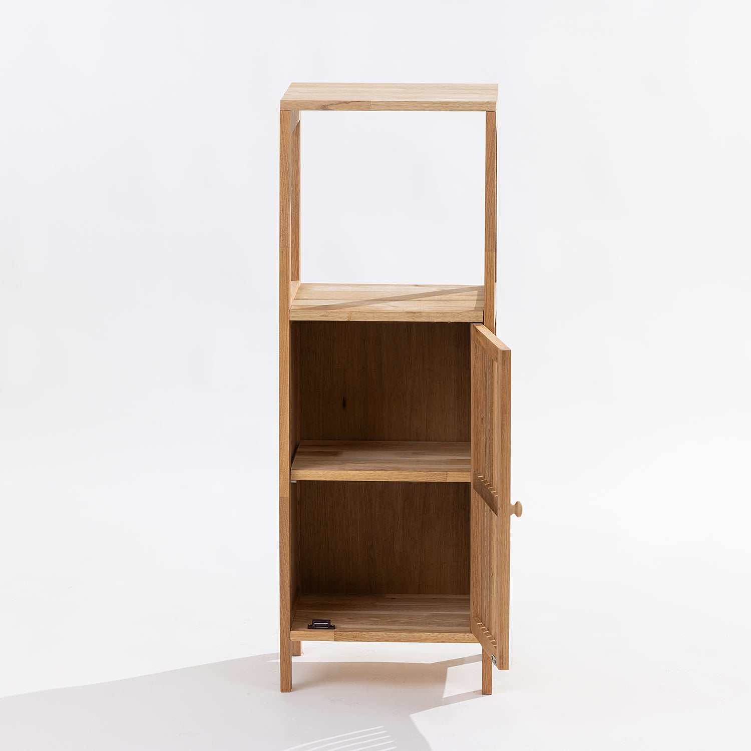 A minimalist wooden storage cabinet with a light natural wood finish, featuring three open shelves and one open door with a small round wooden knob, set against a plain white studio background.