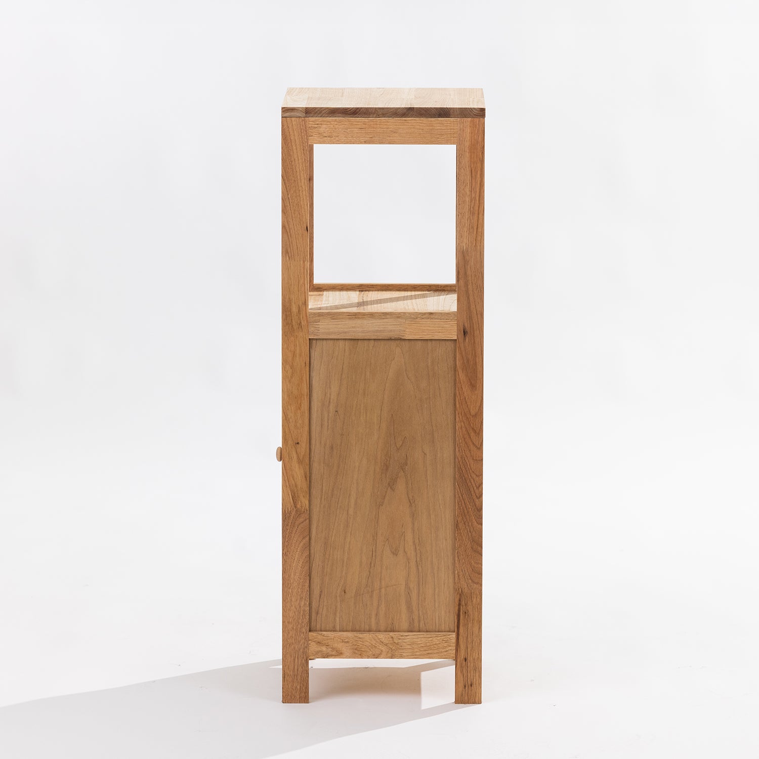 A tall, narrow wooden cabinet or display stand made of warm medium-brown wood, featuring a single open upper shelf and a lower cabinet door with a small round knob, photographed against a clean white studio background. Side View