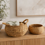 Set of two natural water hyacinth baskets displayed on a wooden console, with the larger handled basket styled with a folded towel and body brush.