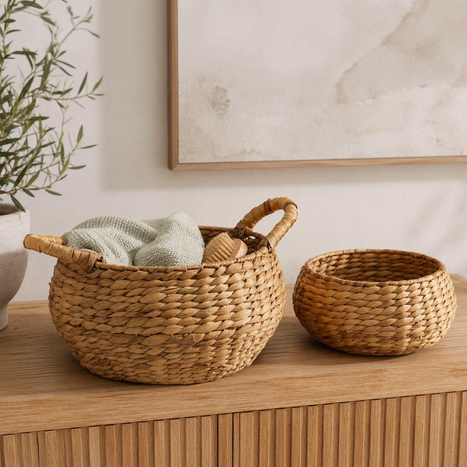 Set of two natural water hyacinth baskets displayed on a wooden console, with the larger handled basket styled with a folded towel and body brush.
