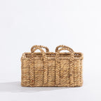 Small natural woven rectangular basket with braided handles, shown against a white background.