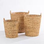 Three woven natural fiber storage baskets in different sizes with top handles, arranged on a white studio surface against a white background.