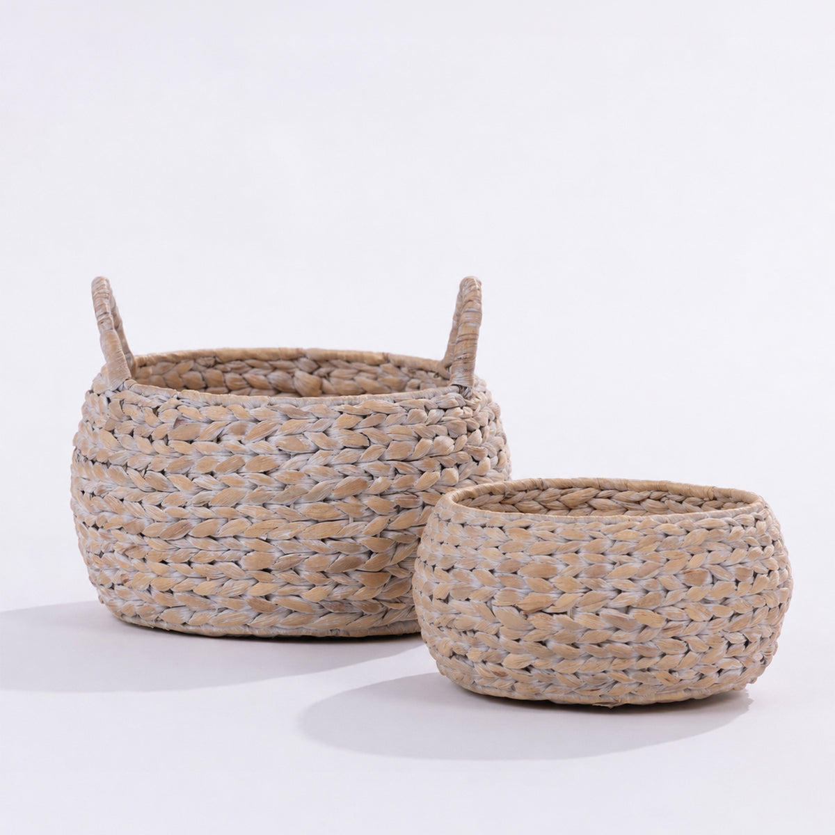 Two round woven baskets made from natural seagrass or straw, one larger with side handles and one smaller, displayed on a clean white studio background.