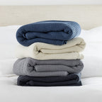 A stylish stack of waffle-weave blankets in blue, ivory, grey, and coal tones adds layered texture and understated elegance to the bedroom setting.