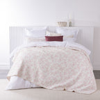 A soft blush floral quilted blanket is layered over crisp white bedding with coordinating cushions, creating a fresh and inviting bedroom look.