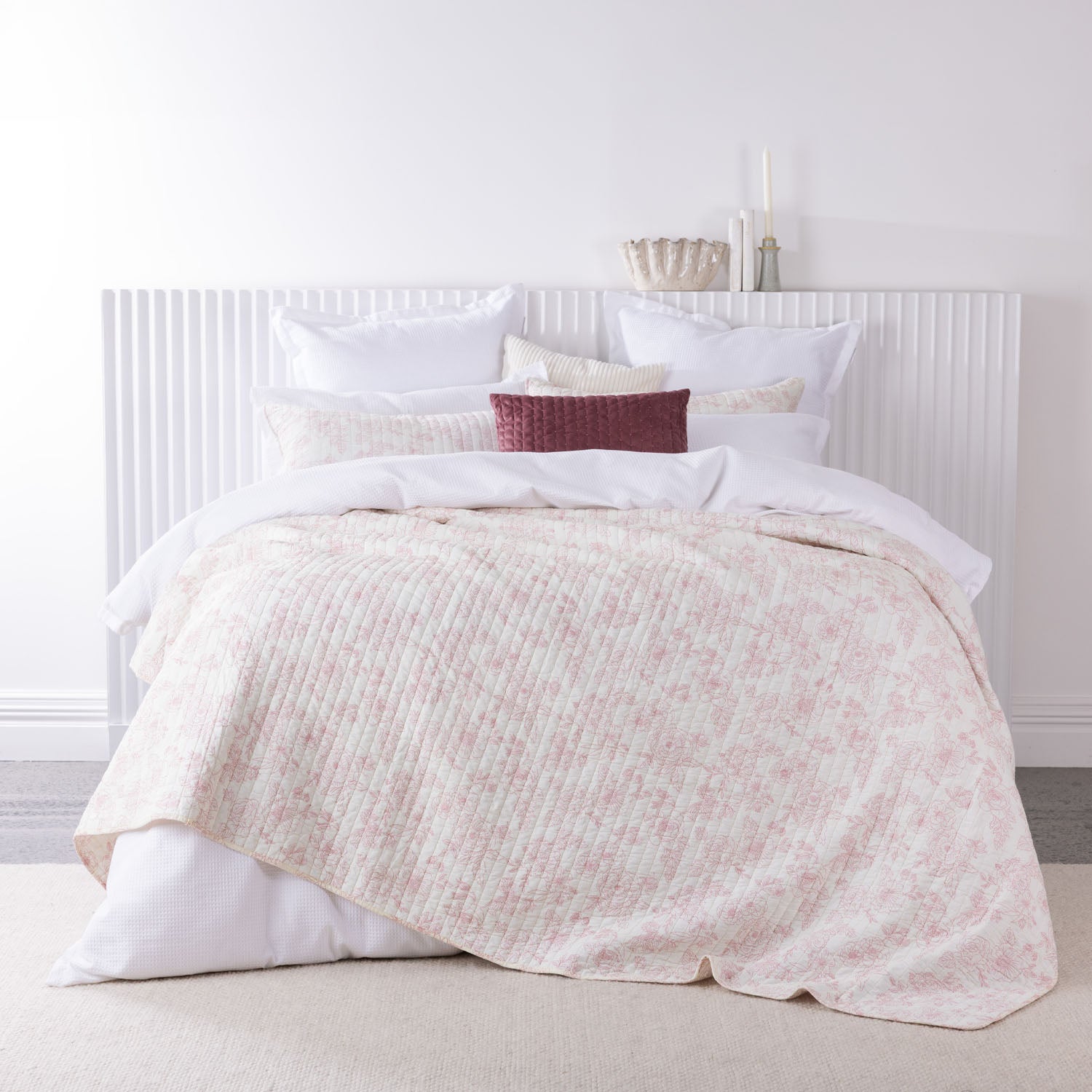A soft blush floral quilted blanket is layered over crisp white bedding with coordinating cushions, creating a fresh and inviting bedroom look.