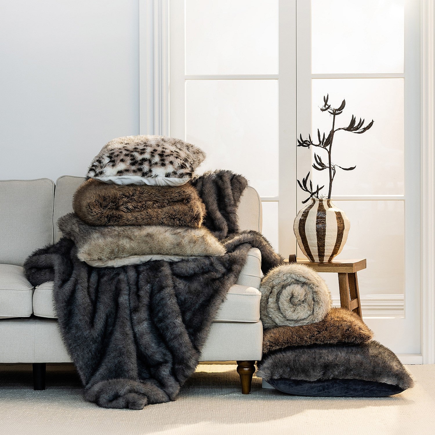 A beautifully styled living room scene featuring layered faux-fur throws, blankets and cushions in rich neutral and animal-inspired tones, creating a warm and luxurious atmosphere.