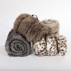 A stylish collection of plush faux-fur blankets in charcoal, taupe, ivory, and snow leopard patterns, elegantly rolled and finished with rich brown leather carry straps.