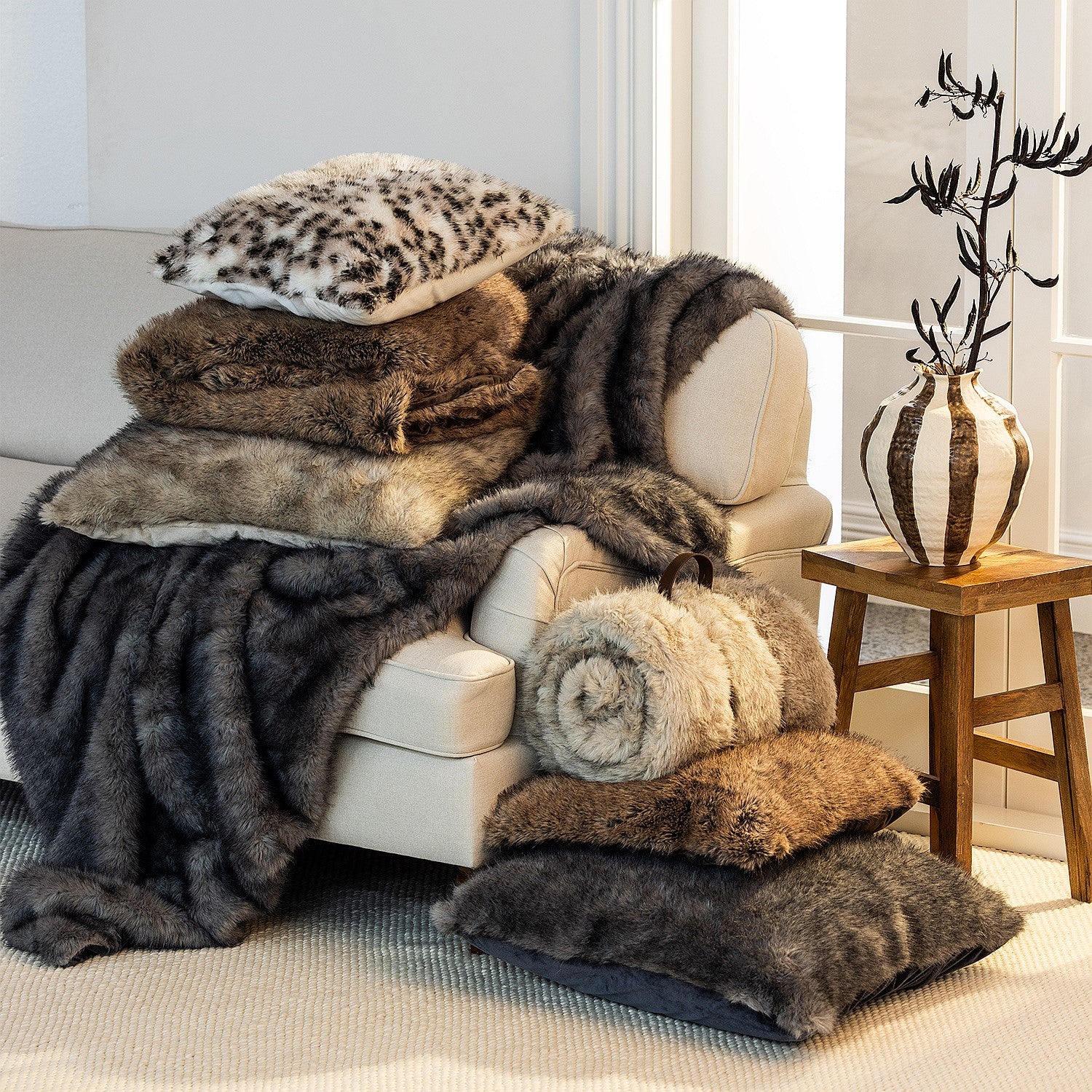 A beautifully styled living room scene featuring layered faux-fur throws, blankets and cushions in rich neutral and animal-inspired tones, creating a warm and luxurious atmosphere.