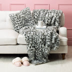 Grey faux fur blanket and cushion set styled on a cream sofa with pink slippers and a mug.