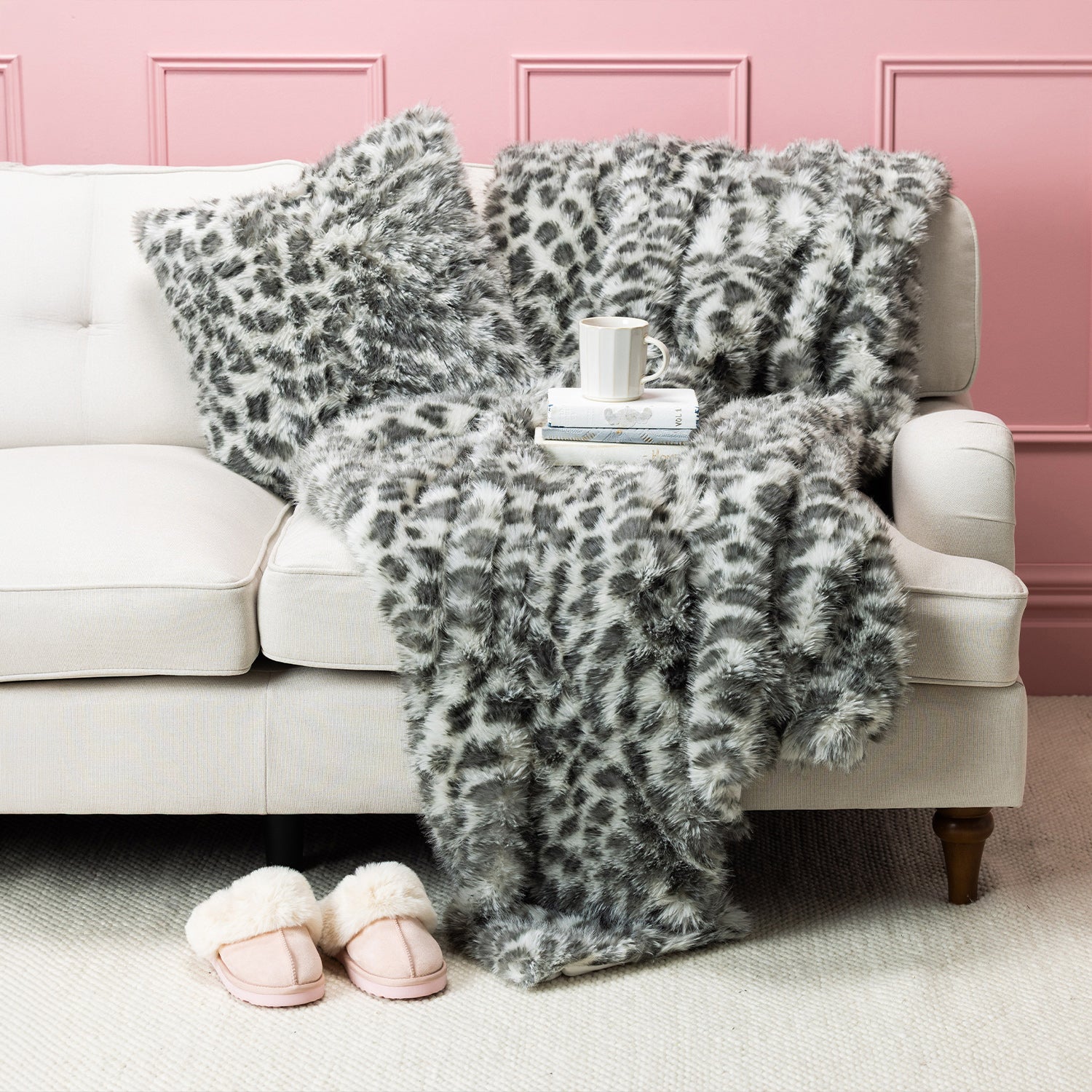 Grey faux fur blanket and cushion set styled on a cream sofa with pink slippers and a mug.