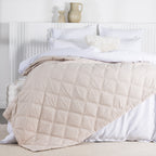A almond milk quilted blanket with a square-stitched pattern is draped across a neatly made white bed in a bright, contemporary bedroom.