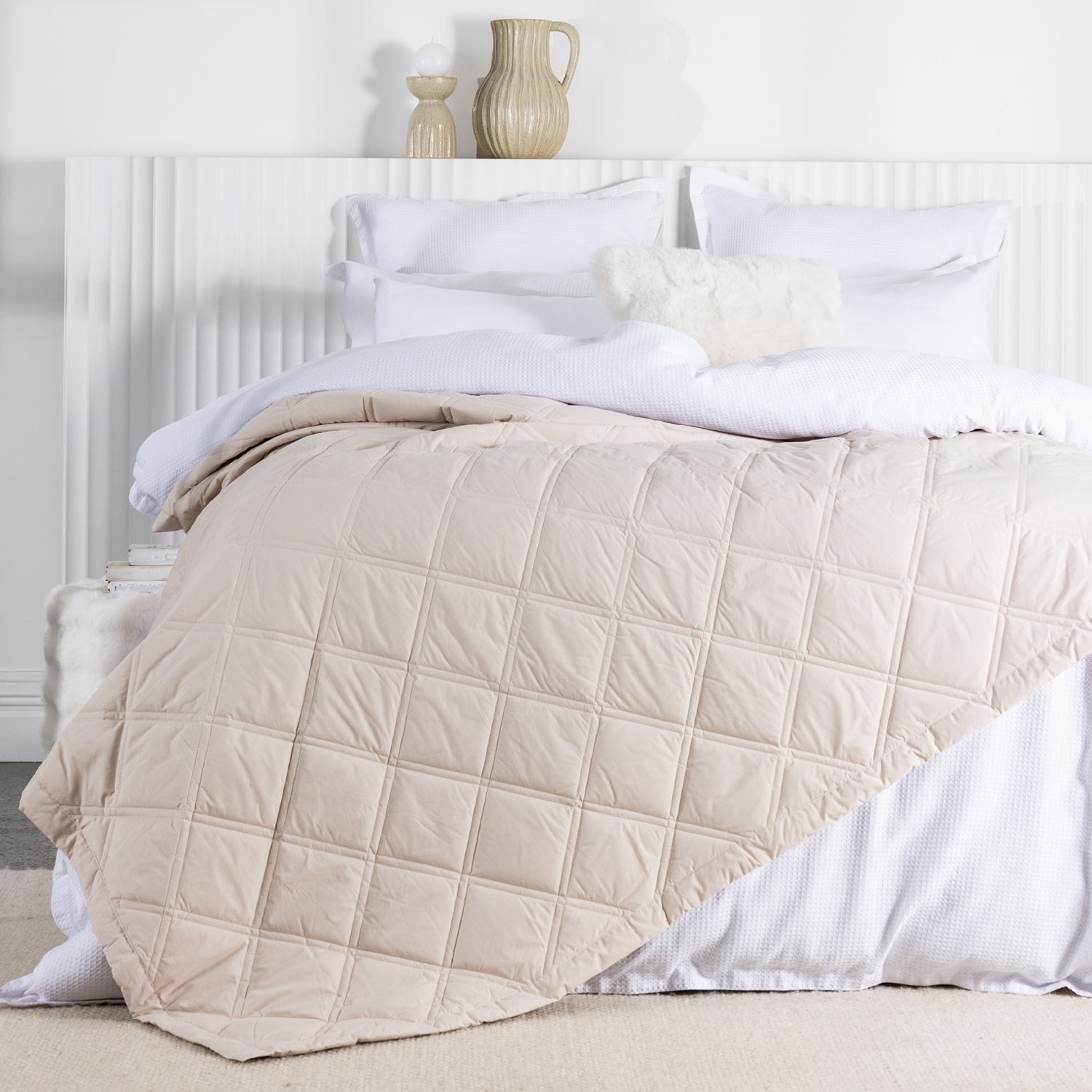 A almond milk quilted blanket with a square-stitched pattern is draped across a neatly made white bed in a bright, contemporary bedroom.