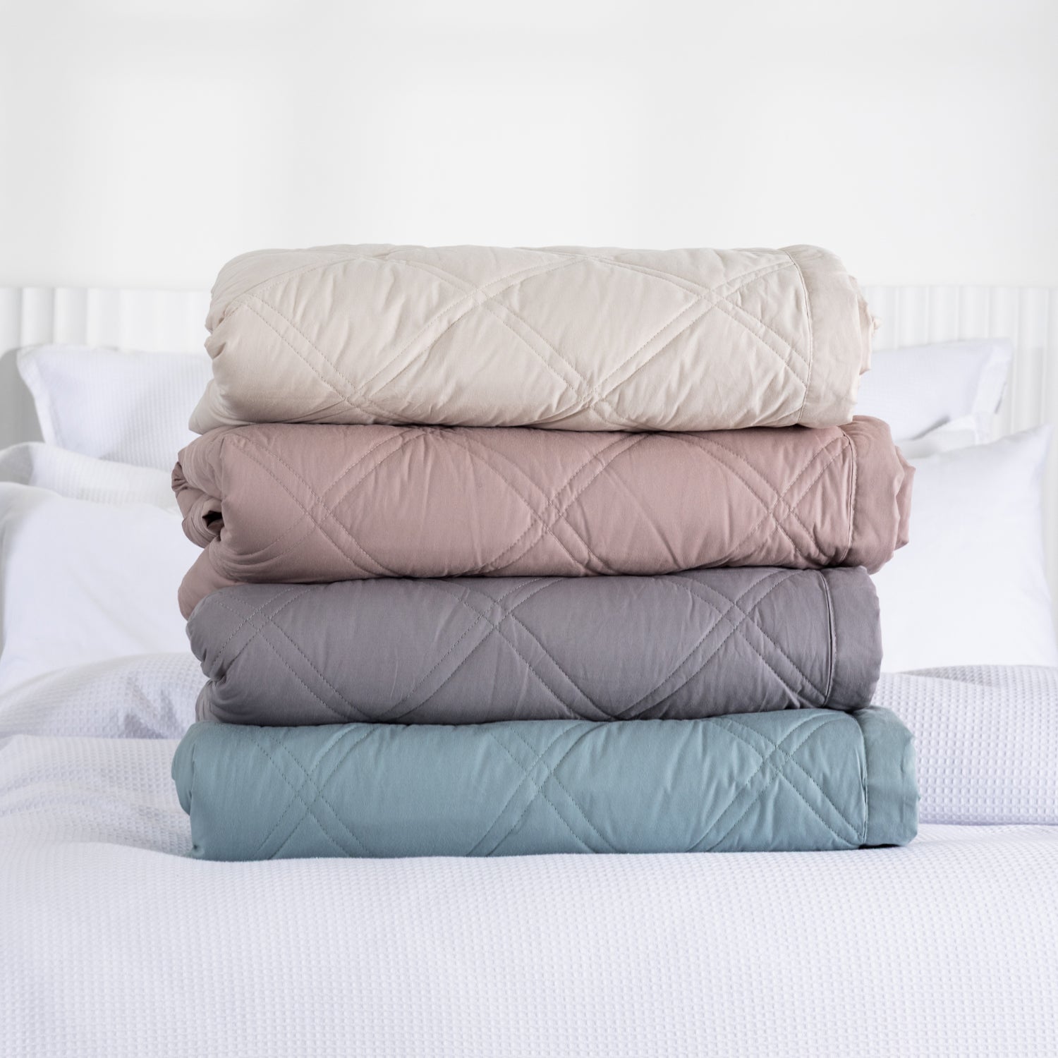 A stack of four folded quilted blankets in almond milk, woodrose, grey, and sage blue sits neatly on a white bed in a bright, minimalist bedroom.