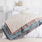 Four quilted blankets in almond milk, woodrose, sage blue, and gray are layered and draped across a neatly made white bed in a bright, minimalist bedroom.