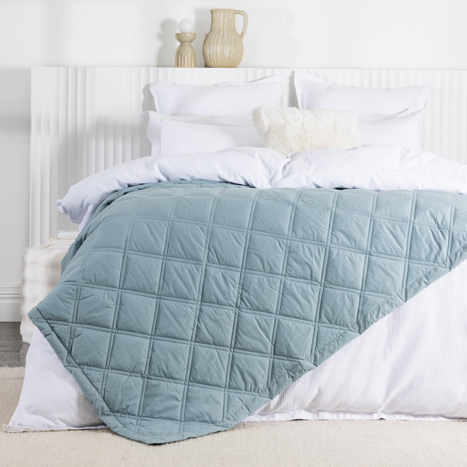 A sage blue quilted blanket with a square-stitched pattern is draped across a neatly made white bed in a bright, contemporary bedroom.