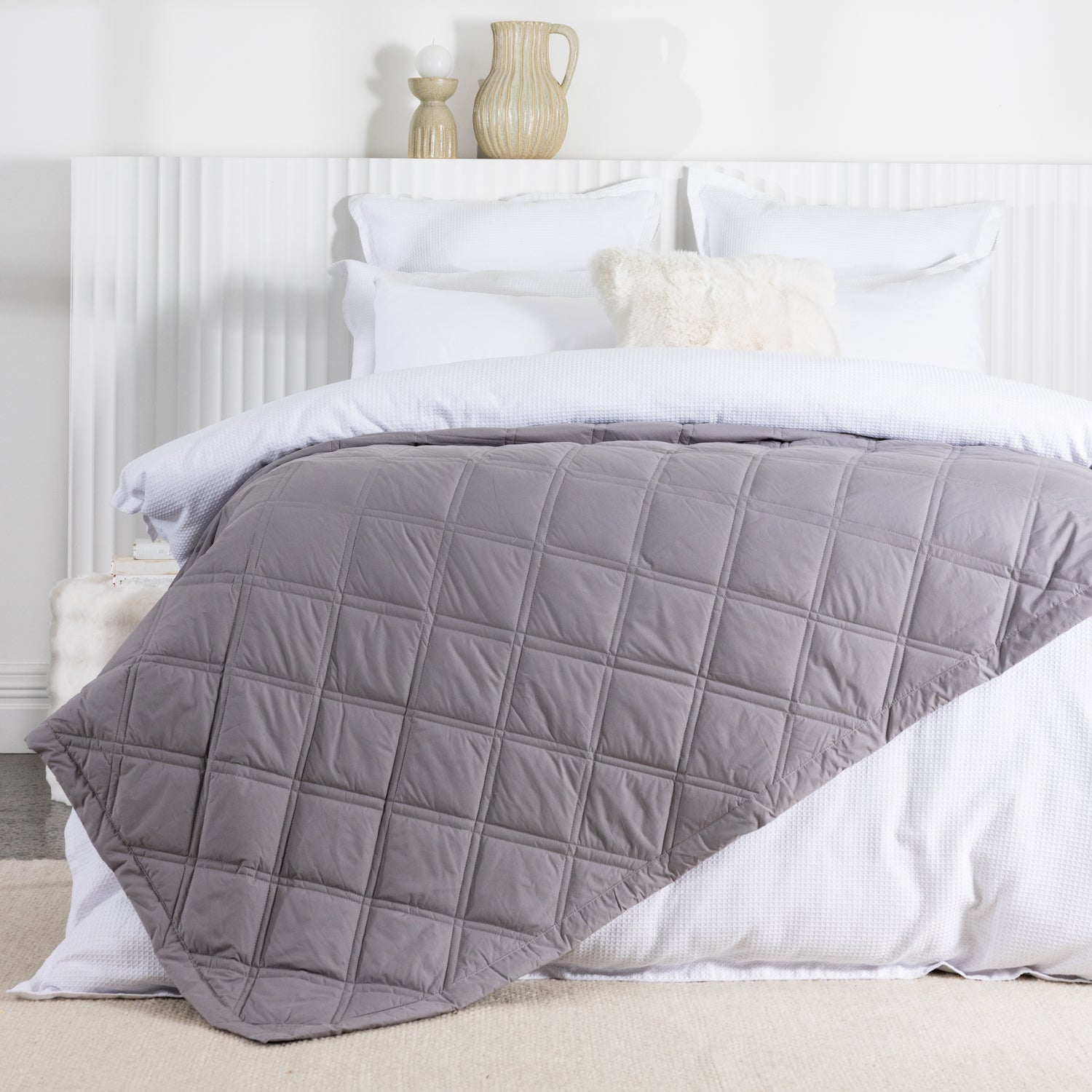 A soft gray quilted blanket with a square-stitched pattern is draped across a neatly made white bed in a bright, contemporary bedroom.
