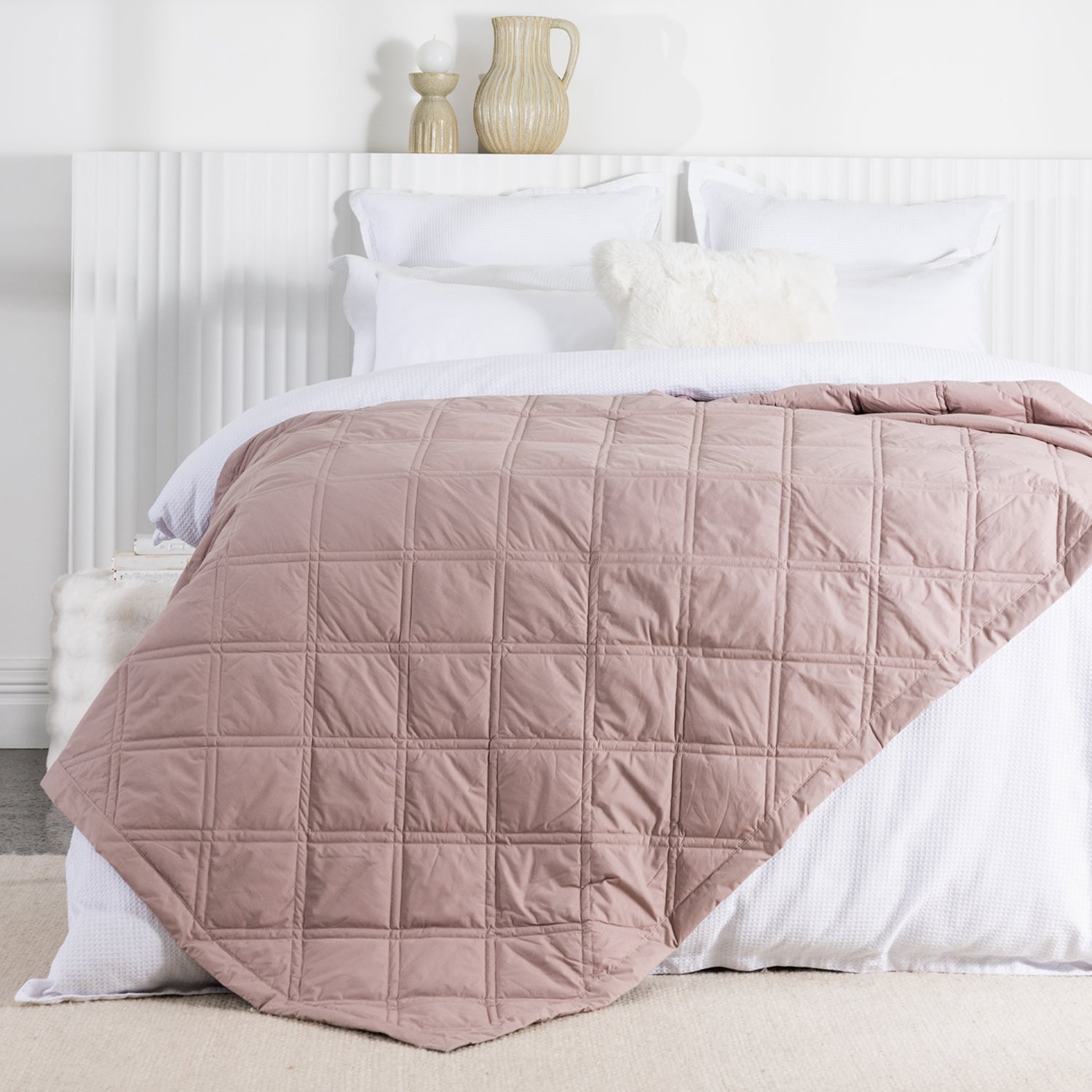 A rosewood quilted blanket with a square-stitched pattern is draped across a neatly made white bed in a bright, contemporary bedroom.