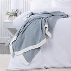 Cove Bayview Muslin Blanket in Denim with a textured weave and contrasting white border, draped across a white bed.