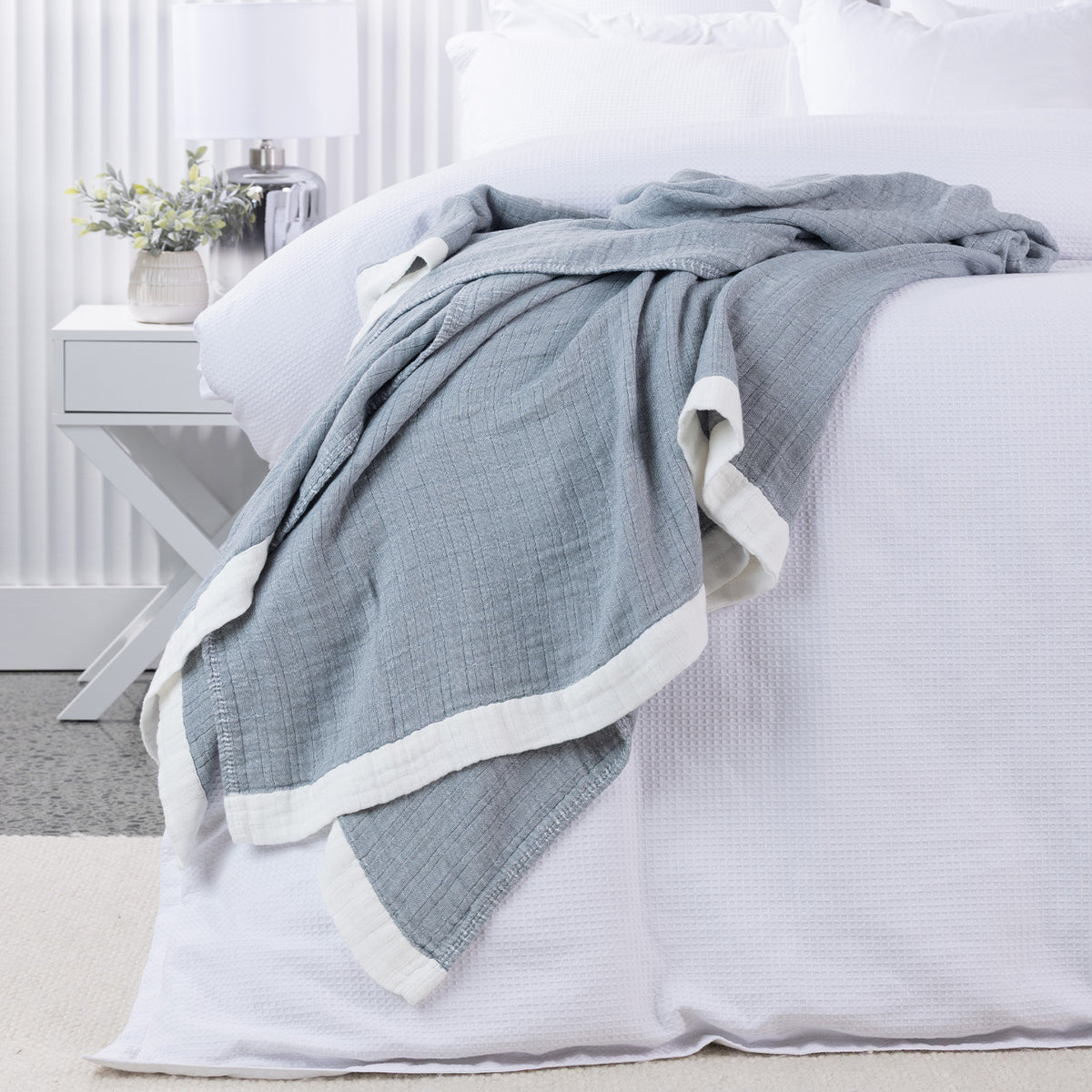 Cove Bayview Muslin Blanket in Denim with a textured weave and contrasting white border, draped across a white bed.