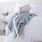 Cove Bayview Muslin Blankets in Natural, Sage and Grey stacked on a white bed, with a Denim blanket draped across the front.