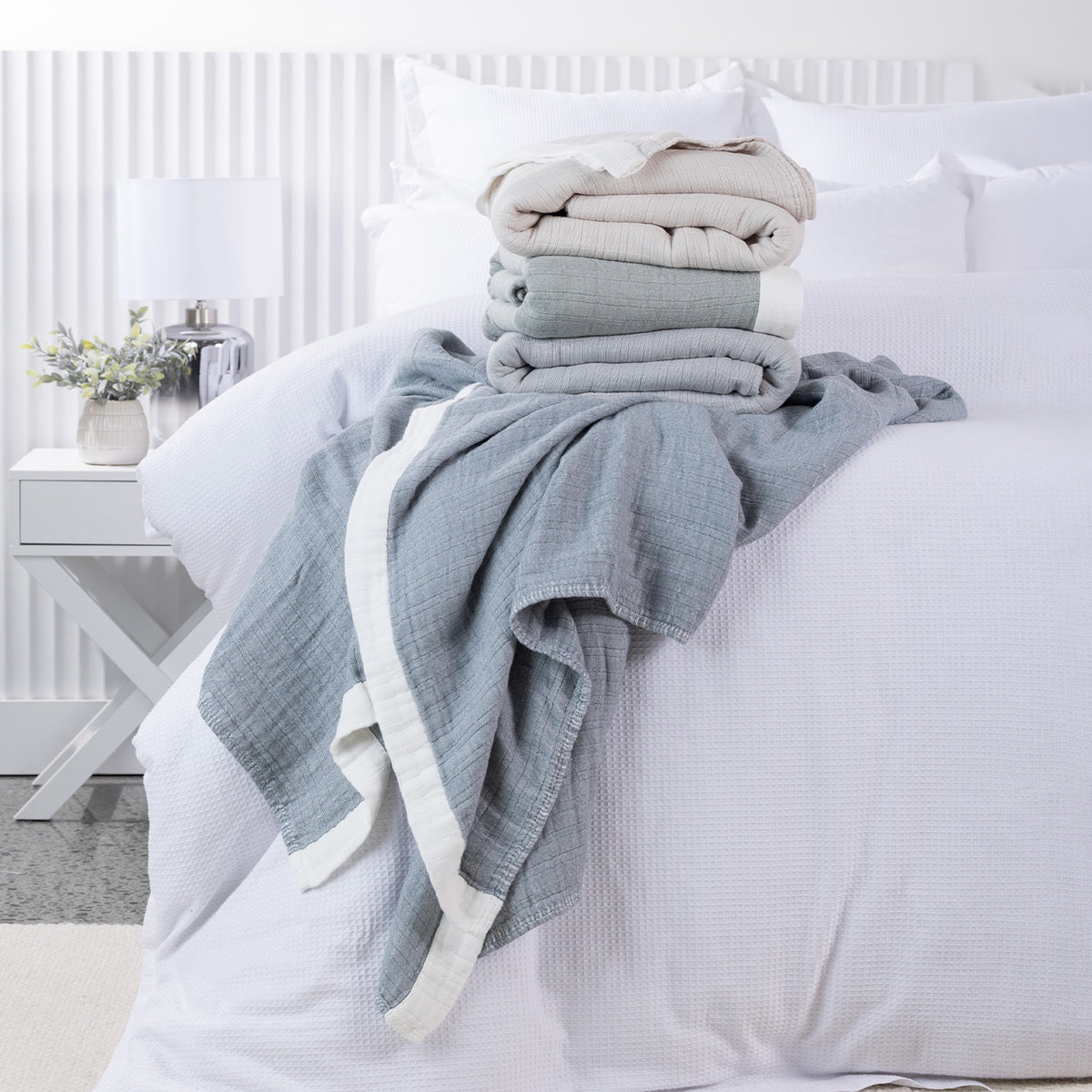 Cove Bayview Muslin Blankets in Natural, Sage and Grey stacked on a white bed, with a Denim blanket draped across the front.